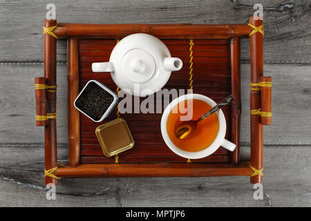 Table Top Blick auf weißer Keramik Teekanne, heiße Tasse schwarzer Tee mit silbernen Löffel und öffnete Dosis mit loser Tee, auf Bambus Tablett serviert auf gr Stockfoto