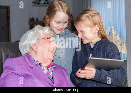 Eine 95-jährige Frau lacht über ihre 7-jährige und 9-jährige Urenkelin. Sie sitzt auf einem Stuhl in Ihrem Wohnzimmer. Die beiden Mädchen sind Stand Stockfoto