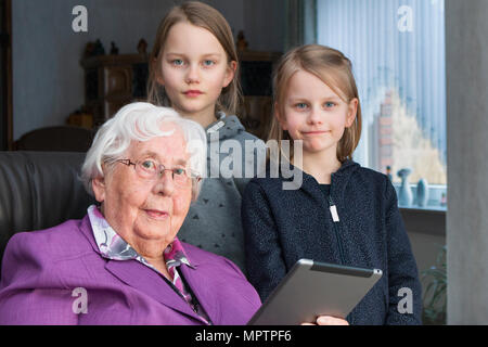 Eine 95-jährige Frau und ihre 7-jährige und 9-jährige Urenkelin Blick in die Kamera. Sie sitzt auf einem Stuhl in Ihrem Wohnzimmer. Die beiden GI Stockfoto