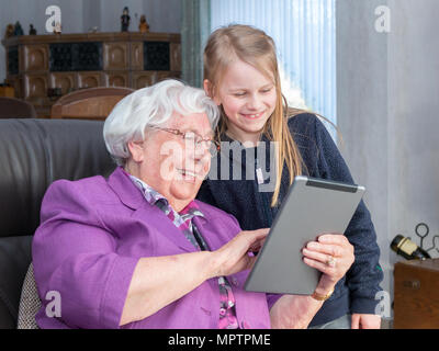 Eine 95-jährige Frau hält ein Tablet und zeigt etwas lustig zu ihrem Enkel. Sie sitzt auf einem Stuhl in Ihrem Wohnzimmer und beide sind auf der Suche Stockfoto