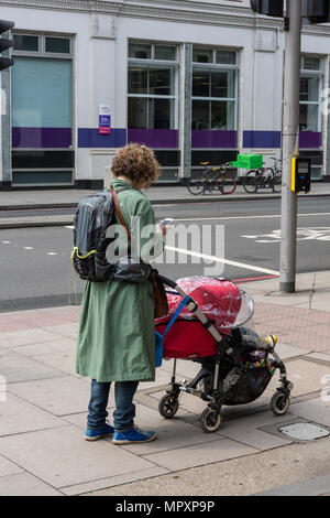 Eine Frau mit einem Kinderwagen mit Kind, während Sie ein Handy an der Seite der Straße, die einen Rucksack und Simsen. Mutter und Kind. Das Bemuttern. Stockfoto