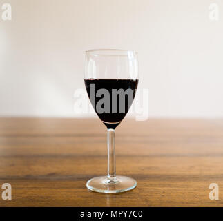 Ein Glas halb voll dunkler Rotwein stehen auf Holz Tisch gegen weiße Wand in verschwommenen Hintergrund. Stockfoto