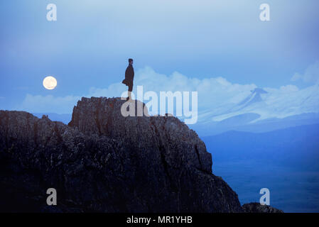 Einsamer Mann am Abend Zeit depressiv steht auf einer Klippe oder Big Rock gegen Berge und Mond Stockfoto
