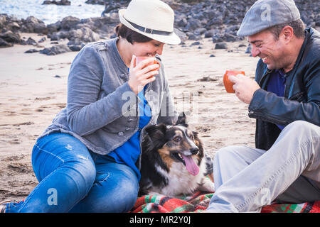 Schöne Paar mittleren Alters in der Freizeit am Strand im Urlaub im Sommer mit einem süßen Haustier Border Collie. Alle zusammen mit einer Tasse Kaffee zu anejoy Stockfoto