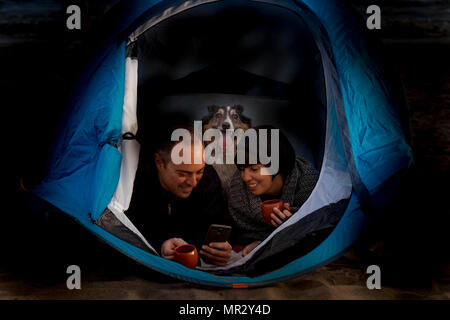 Camping alternative Ferienhäuser Lebensstil mit Paar mittleren Alters mit Border Collie Hund in einem Zelt in der Nacht. Beide trinken Tee oder Kaffee und die m prüfen Stockfoto