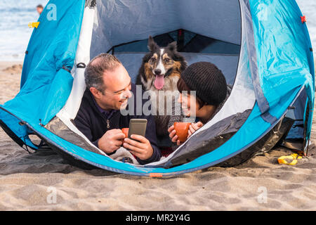 Schön Schön paople mit niedlichen Welpen in einem Zelt mit Mobiltelefon Nachricht senden möchten, oder nehmen Sie ein Bild auf. Der Strand camping Freiheit alternative Ferienhäuser i Stockfoto