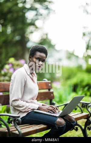 Afro-amerikanische Frauen, die sie mit dem Laptop sitzt auf der Holzbank. Freundliche nette schwarze Mädchen auf der Parkbank mit dem Netbook. Stockfoto