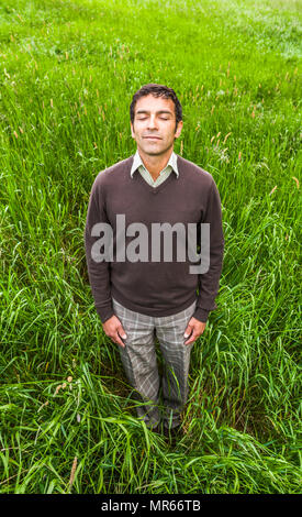 Ein Mann stand in einer grünen Wiese meditieren mit geschlossenen Augen. Stockfoto