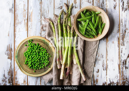 Vielzahl von Roh ungekocht organische Junge grüne Gemüse, Spargel, Erbsen, pod Erbse in keramischen Platten auf Leinen über weiß Holzbrett Hintergrund. T Stockfoto