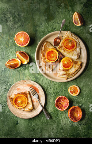 Hausgemachte crepes Pfannkuchen in keramischen Platten mit blutigen Orangen und Rosmarin Sirup mit in Scheiben geschnittenen sizilianischen Rotwein Orangen über grüne Textur backgroun serviert. Stockfoto