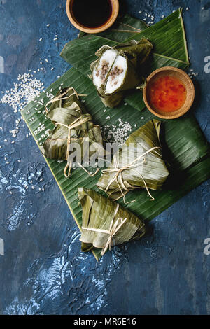 Asiatische Reis piramidal gedämpft Knödel aus Tapioka Mehl von Reis mit Fleisch füllen in Bananenblättern. Zutaten und Saucen oben über blaue Textur zurück Stockfoto