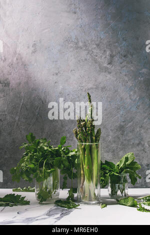 Roh ungekocht organischen grünen Spargel, Bündel von koriander Kräuter und frischer Minze in Gläsern auf einem weißen Tisch mit grau Struktur Wand als backgroun Stockfoto