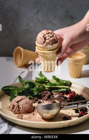 Die Hand des Kindes nehmen Sie hausgemachte Schokolade Himbeere Eis in kleinen Waffel Cup. Gefrorene Beeren, frische Minze, dunkle Schokolade und Metalllöffel in ce-gehackt Stockfoto