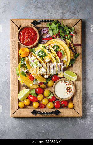 Auswahl an vegetarischen Mais Tacos mit Gemüse, Salat, Chili Pfeffer serviert auf Holz- Fach mit Tomaten und Sahne Saucen mit Zutaten oben Stockfoto