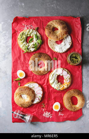 Auswahl an hausgemachten Bagels mit Sesam, Frischkäse, Pesto, Eier, Rettich, Kräutern auf Rot zerknittertes Papier mit Zutaten oben serviert über Stockfoto