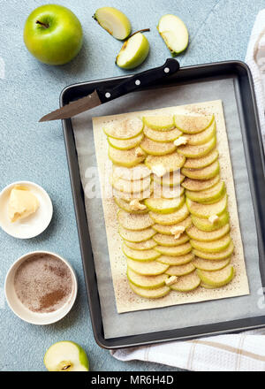 Eine ungekochte rechteckige Apfelkuchen auf ein Backblech mit einer Schüssel mit Zimt, Zucker und einer Schüssel Butter. Stockfoto