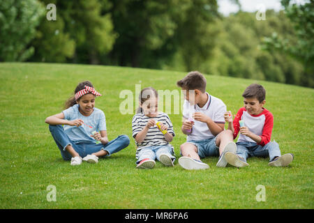 Happy multiethnischen Kinder spielen mit Seifenblasen beim Sitzen auf der grünen Wiese im Park Stockfoto