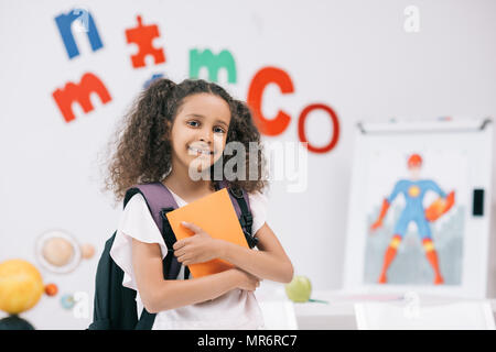 Adorable african american Schulmädchen mit Rucksack holding Lehrbuch und lächelnd an der Kamera im Klassenzimmer Stockfoto