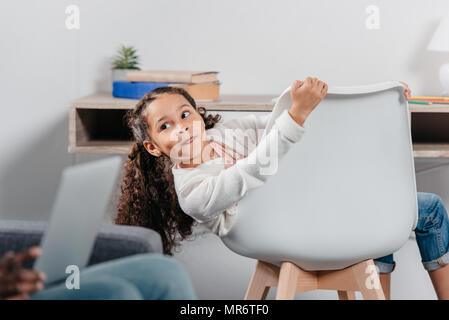 Adorable african american Mädchen sitzt auf einem Stuhl und Blick auf Vater sitzt mit Laptop auf den Knien Stockfoto