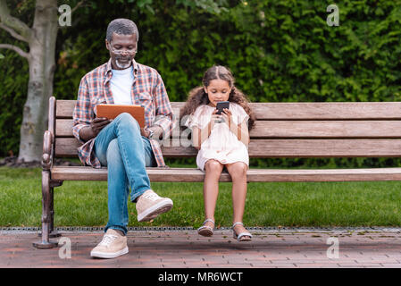 Adorable African American Girl und ihr Großvater sie ihr Smartphone und digitale tablet, während auf der Bank im Park sitzen Stockfoto
