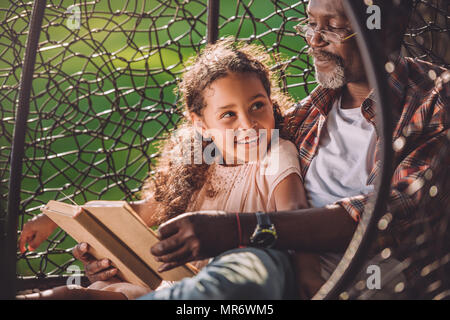 Lächelnd afrikanische amerikanische Enkelin lesen Buch während in Schwingen Hängesessel mit Großvater sitzen Stockfoto