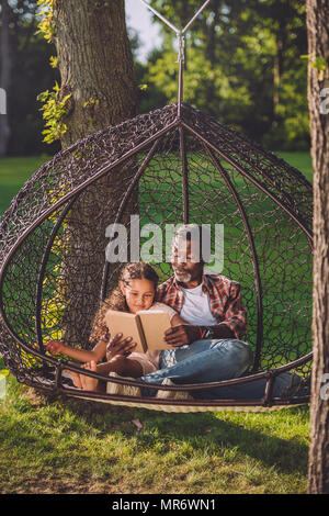 Kleine afrikanische amerikanische Enkelin lesen Buch während in Schwingen Hängesessel mit Großvater sitzen Stockfoto