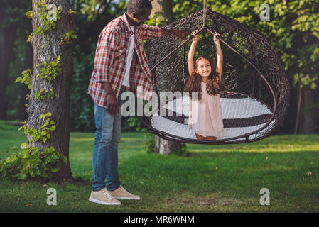 African American Girl schwingen auf Hängesessel, während ihr Großvater in der Nähe von im Park Stockfoto
