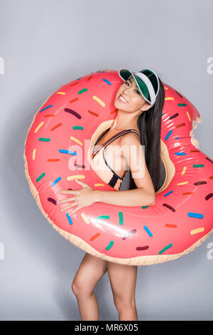 Halbe Länge geschossen von lächelnden Asiatin in Coral Badeanzug und Visor, trug einen Donut pool schwimmen um ihren Körper und Kamera Stockfoto