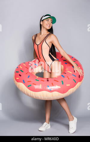 In voller Länge geschossen von fröhlichen asiatische Frau in Coral Badeanzug und Visor, trug einen Donut pool schwimmen um ihren Körper Stockfoto