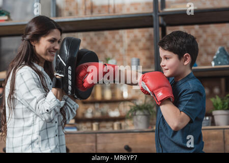 Little Boy in Boxhandschuhe treten eines Stanzen Pad von seiner Mutter gehalten Stockfoto