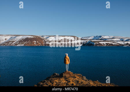 Rückansicht der jungen Frau, die auf der felsigen Küste und Suchen an der majestätischen isländische Landschaft mit Golf und die schneebedeckten Berge Stockfoto