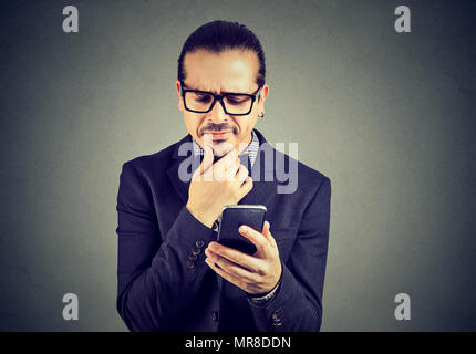 Business Mann in Brillen ratlos und skeptisch auf der Suche während der Verwendung von Handys auf grauem Hintergrund. Stockfoto