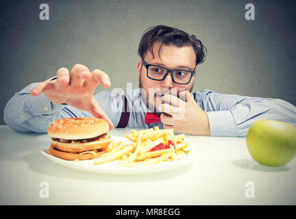 Junge Mann in Gläser, die cravings für Cheeseburger mit Pommes anstelle von gesunden grünen Apfel. Stockfoto