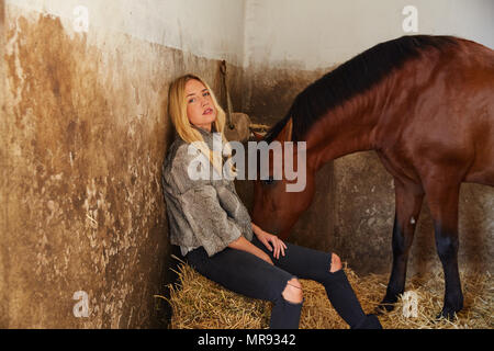 Blonde Frau auf einem stabil mit einem braunen Pferd auf Stroh ballen Stockfoto