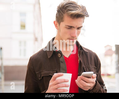 Junger Mann Kaffee in der Tasse und suchen ernsthafte beim Surfen Smartphone und Lesen schlechte Nachrichten außerhalb. Stockfoto