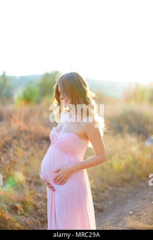 Schwangere Frau umarmen Bauch, tragen rosa Kleid in der Steppe mit Sonnenstrahlen Hintergrund. Stockfoto