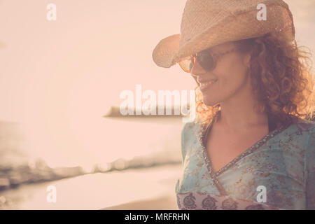 Schöne und wunderbare blondes lockiges Haar Modell mittleren Alters mit Cowboyhut Lächeln und am Strand mit goldenen Sonnenuntergang im Hintergrund darstellen. Farbige ein Stockfoto