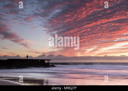 Ein Mann steht auf einer verfallenden Jetty in Ehrfurcht vor einem lebendigen Sonnenuntergang, erzeugen einen starken Kontrast zwischen den städtischen Santa Monica Bay und natürliche Schönheit in Los Ein Stockfoto