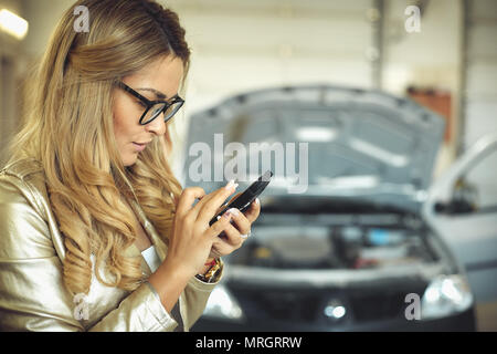 Schöne junge blonde Frau in Gläsern ruft das Telefon auf und Automechaniker für die automatische Reparatur Stockfoto