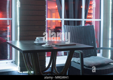 Tisch und Stuhl in der Nacht Cafe mit einem Becher aus Kaffee und das Konto vor dem Hintergrund eines Fensters Stockfoto