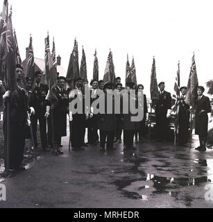 1964, historische, Veteran Soldaten der britischen Streitkräfte mit Regimental flags Line-up Sammeln draußen auf einer nassen Straße vor einem Erinnerung Sonntag März für die Streitkräfte, Nächstenliebe, die Royal British Legion, Bucks, England, UK. Stockfoto