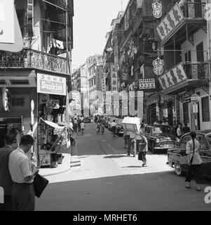 1950, historische Ansicht einer hektischen Straße in Hongkong, Asien, mit Fahrzeugen an der Seite und Geschäften und Wohnungen oben geparkt, die 'multi-Geschichte" Gebäude. Es blieb eine Britische Kolonie nach WW2 und sah einen großen Zustrom von Einwanderern aus den Nachbarländern China, die die Bevölkerung und Kick erhöht - das rasante wirtschaftliche Wachstum gestartet. Stockfoto