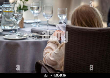 Das Kind sitzt auf einem Stuhl am festlich gedeckten Tisch und liest eine Broschüre. Stockfoto