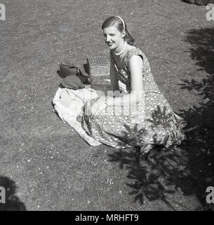 1950er Jahre, historische Bild zeigt eine elegante Dame in einem langen Kleid mit pear Halskette draußen auf dem Rasen mit Ihren Picknickkorb, England, UK. Stockfoto