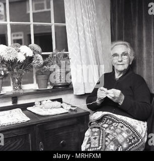 1960, historische, alte Dame, der in den geschützten Unterkunft, neben einem Fenster Kitting, England, Großbritannien sitzen. Stockfoto
