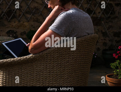 Seitenansicht Profil der braungebrannte reife Frau mit Brille in Rattan Gartenstuhl sitzen auf der Terrasse mit den Blumentöpfen auf dem Bildschirm Stockfoto