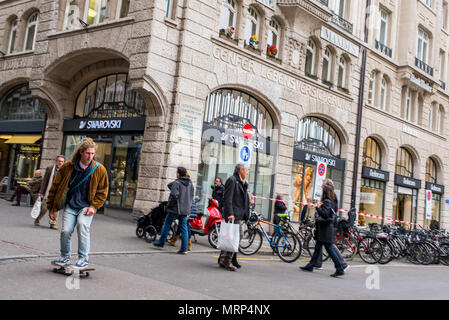 Junge männliche Kursteilnehmer mit langen Haaren skateboarding in einer belebten Straße Freie Strasse in Basel City Center unter Menschen zu Fuß Stockfoto