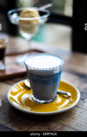 Heiße Milch mit Butterfly pea gemischt und Milchschaum auf der Oberseite aus Glas, mit gelben Festplatte auf hölzernen Tisch im Cafe Shop Stimmung und Ton schmücken, selektive Stockfoto