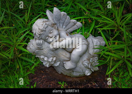 Kleiner Engel schläft. Skulptur eines kleinen Engel schlafen auf Baum einen Ast. Es liegt auf einem alten Baumstumpf mit grünen Blättern umgeben. Stockfoto