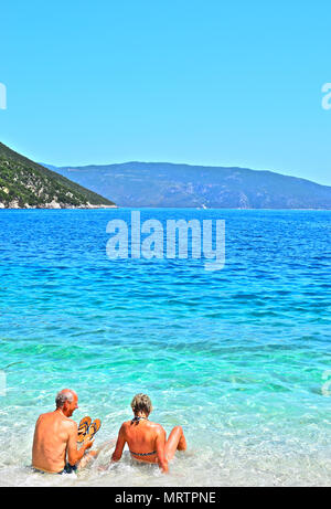 Ein älteres Paar Spaß und genießen Sie im kristallklaren Wasser des Ionischen Meeres an Antisamos Beach in der Nähe von Sami, Kefalonia, Griechenland sitzen. Stockfoto